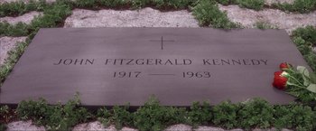 Movie still from “JFK” (1991), directed by Oliver Stone – A grave marker with a cross on it in the grass; Wide shot, High angle