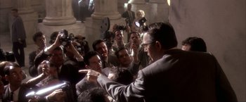 Movie still from “JFK” (1991), directed by Oliver Stone – A group of people gathered around a man in a gray suit; Medium shot, Over the shoulder angle