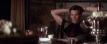 Movie still from “JFK” (1991), directed by Oliver Stone – A man sitting at a dinner table with his hand behind his head; Medium shot, Over the shoulder angle