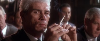 Movie still from “JFK” (1991), directed by Oliver Stone – A man in a suit and tie smoking a cigarette in front of another man in a suit and tie; Close Up shot, Low angle