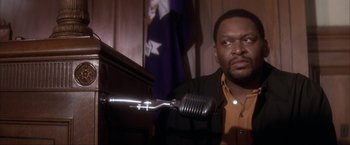 Movie still from “JFK” (1991), directed by Oliver Stone – A man holding a microphone in front of a microphone stand; Close Up shot, Over the shoulder angle