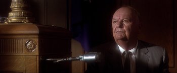 Movie still from “JFK” (1991), directed by Oliver Stone – An older man wearing a suit and tie with a microphone in front of him; Close Up shot, Low angle