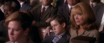 Movie still from “JFK” (1991), directed by Oliver Stone – A boy in a suit and bow tie sitting in front of a group of people; Close Up shot, Over the shoulder angle