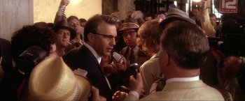 Movie still from “JFK” (1991), directed by Oliver Stone – A group of people gathered around a man in a suit and tie; Close Up shot, Over the shoulder angle