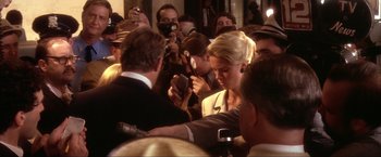 Movie still from “JFK” (1991), directed by Oliver Stone – A crowd of people gathered around a man holding up a cell phone; Medium shot, Over the shoulder angle