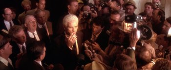 Movie still from “JFK” (1991), directed by Oliver Stone – A man smoking a cigarette in front of a group of people; Medium shot, High angle
