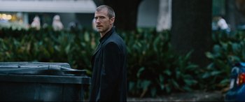 Movie still from “Jack Reacher: Never Go Back” (2016), directed by Edward Zwick – A man standing in front of bushes and a trash can; Medium shot, Over the shoulder angle