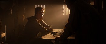 Movie still from “Jack the Giant Slayer” (2013), directed by Bryan Singer – A man sitting at a wooden table in a dimly - lit room; Medium shot, Over the shoulder angle