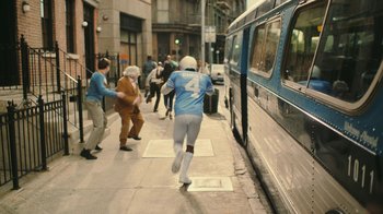 Movie still from “Jackass Forever” (2022), directed by Jeff Tremaine – A man running down the street in front of a bus; Wide shot, High angle