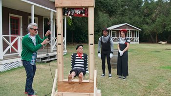 Movie still from “Jackass Forever” (2022), directed by Jeff Tremaine – A group of people standing around a guillotine in the grass; Wide shot, Over the shoulder angle