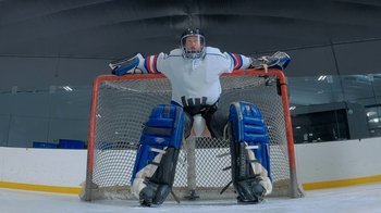Movie still from “Jackass Forever” (2022), directed by Jeff Tremaine – A man in a white and blue hockey uniform is sitting on a goal net; Wide shot, Low angle