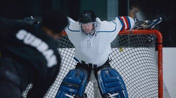 Movie still from “Jackass Forever” (2022), directed by Jeff Tremaine – A hockey player in a white uniform and blue pads; Medium shot, Low angle