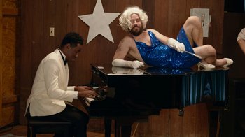 Movie still from “Jackass Forever” (2022), directed by Jeff Tremaine – A man sitting on top of a piano next to another man; Medium shot, Low angle