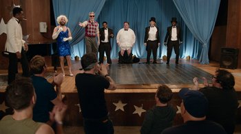 Movie still from “Jackass Forever” (2022), directed by Jeff Tremaine – A group of people on a stage in front of an audience; Wide shot, Over the shoulder angle