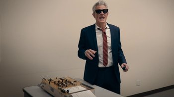 Movie still from “Jackass Forever” (2022), directed by Jeff Tremaine – A man in a suit and tie standing next to a table; Medium shot, Low angle