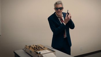 Movie still from “Jackass Forever” (2022), directed by Jeff Tremaine – A man standing next to a table with a cell phone in front of him; Medium shot, High angle
