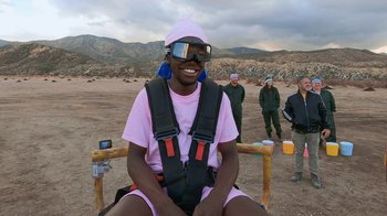 Movie still from “Jackass Forever” (2022), directed by Jeff Tremaine – A man sitting on a chair wearing a pink shirt and a hat; Medium shot, High angle