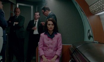 Movie still from “Jackie” (2016), directed by Pablo Larraín – A woman in a pink suit sitting on a chair in front of two men; Medium shot, Over the shoulder angle