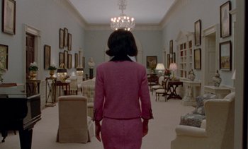 Movie still from “Jackie” (2016), directed by Pablo Larraín – A woman in a pink suit standing in a room full of furniture; Wide shot, Low angle