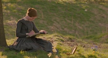 Movie still from “Jane Eyre” (2011), directed by Cary Joji Fukunaga – A woman sitting in the grass writing on a book; Medium shot, High angle