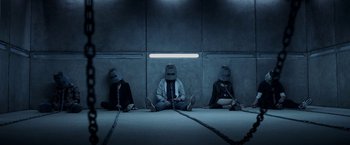 Movie still from “Jigsaw” (2017), directed by Michael Spierig – A group of people sitting on the ground wearing masks; Wide shot, Overhead angle