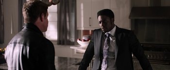 Movie still from “Jigsaw” (2017), directed by Michael Spierig – A man wearing a suit and tie sitting in a kitchen; Medium shot, Over the shoulder angle