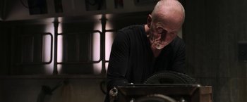 Movie still from “Jigsaw” (2017), directed by Michael Spierig – An older man in a black shirt is looking at a machine; Medium shot, Low angle