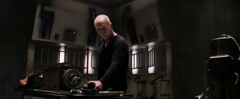 Movie still from “Jigsaw” (2017), directed by Michael Spierig – A bald man in a black shirt is working on a machine; Medium shot, Low angle