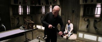 Movie still from “Jigsaw” (2017), directed by Michael Spierig – A man holding a gun while standing next to another man sitting down; Medium shot, Over the shoulder angle