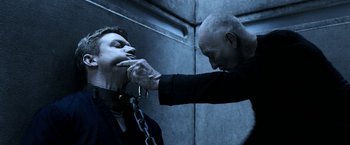 Movie still from “Jigsaw” (2017), directed by Michael Spierig – A man with a chain around his neck is being whipped by another man; Close Up shot, Low angle