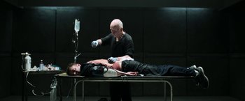 Movie still from “Jigsaw” (2017), directed by Michael Spierig – A person is lying on a table and a man is standing over him; Medium shot, Overhead angle