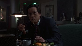 Movie still from “Johnny Mnemonic” (1995), directed by Robert Longo – A man sitting at a dinner table eating food; Medium shot, Low angle