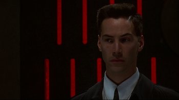 Movie still from “Johnny Mnemonic” (1995), directed by Robert Longo – A man in a suit and tie standing in front of a wall; Close Up shot, Over the shoulder angle