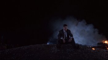 Movie still from “Johnny Mnemonic” (1995), directed by Robert Longo – A man sitting on the ground in front of smoke; Wide shot, Low angle