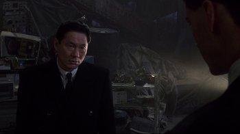 Movie still from “Johnny Mnemonic” (1995), directed by Robert Longo – A man wearing a suit and tie standing in a dark room; Close Up shot, Over the shoulder angle