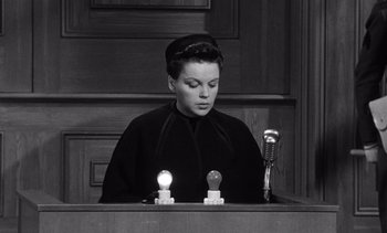 Movie still from “Judgment at Nuremberg” (1961), directed by Stanley Kramer – A woman in a black dress is sitting in front of a microphone and two light bulbs; Medium shot, High angle