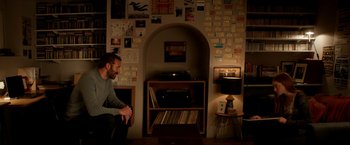 Movie still from “Juliet, Naked” (2018), directed by Jesse Peretz – A man sitting in front of a record player in a room; Wide shot, Over the shoulder angle