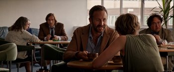 Movie still from “Juliet, Naked” (2018), directed by Jesse Peretz – A man sitting at a table in a room with two other people; Medium shot, Over the shoulder angle