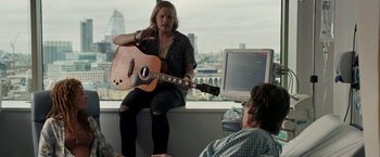 Movie still from “Juliet, Naked” (2018), directed by Jesse Peretz – A man sitting on a windowsill holding a guitar next to another man; Medium shot, Over the shoulder angle