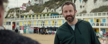 Movie still from “Juliet, Naked” (2018), directed by Jesse Peretz – A man standing on the beach in front of a building; Close Up shot, Over the shoulder angle