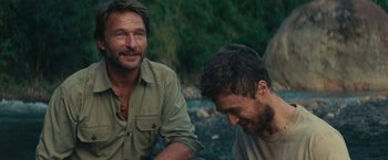 Movie still from “Jungle” (2017), directed by Greg McLean – Two men sitting next to each other near a body of water; Medium shot, Over the shoulder angle