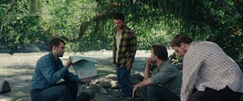 Movie still from “Jungle” (2017), directed by Greg McLean – Two men sitting under a tree near a body of water; Wide shot, Over the shoulder angle