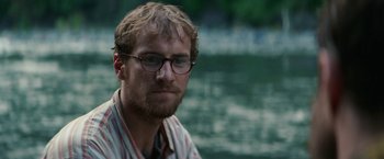 Movie still from “Jungle” (2017), directed by Greg McLean – A person wearing glasses near a body of water; Close Up shot, Low angle