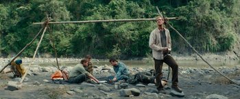 Movie still from “Jungle” (2017), directed by Greg McLean – A group of men sitting on the ground next to a body of water; Wide shot, Low angle