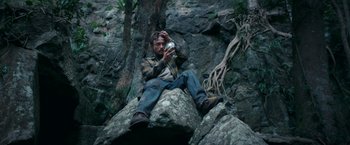 Movie still from “Jungle” (2017), directed by Greg McLean – A man sitting on top of a rock holding a cell phone; Wide shot, High angle