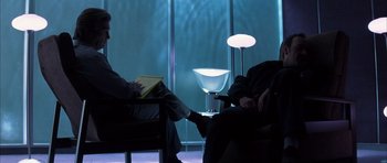 Movie still from “K-PAX” (2001), created by Robert Cochran – Two people sitting in chairs in front of a glass wall; Medium shot, Over the shoulder angle