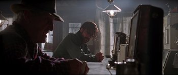 Movie still from “Kalifornia” (1993), directed by Dominic Sena – A man sitting at a table in a dark room; Medium shot, Over the shoulder angle