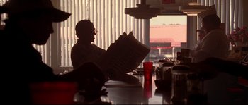 Movie still from “Kalifornia” (1993), directed by Dominic Sena – A man sitting at a table reading a newspaper; Medium shot, Over the shoulder angle