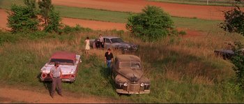 Movie still from “Kalifornia” (1993), directed by Dominic Sena – An old car is parked in a field with people; Extreme Wide shot, High angle