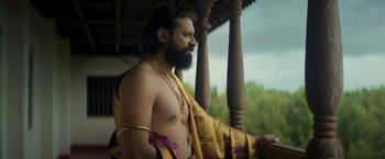 Movie still from “Kantara” (2022), directed by Rishab Shetty – A man with long hair and a beard is wearing a necklace; Medium shot, Low angle